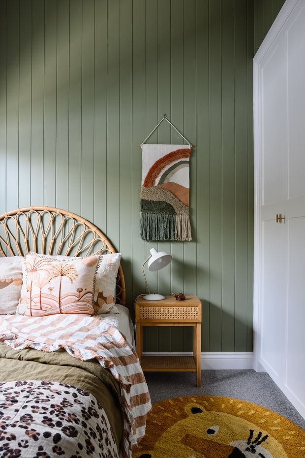 A cozy, compact bedroom with muted sage-green vertical wall panelling, a white paneled built-in wardrobe, and a rattan bedhead. Layered patterned bedding, a small cane-front timber bedside table, a white angled lamp, woven wall hanging, and a playful mustard area rug create a warm, eclectic look.