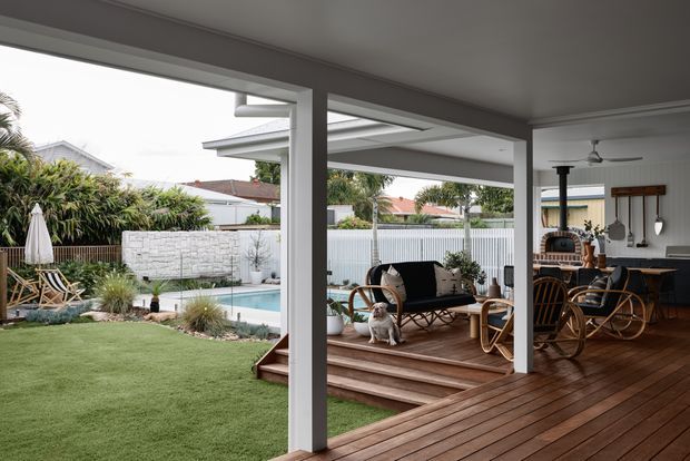 A covered outdoor living and dining area opening onto a landscaped backyard with a small swimming pool. The space features timber decking, white structural posts and ceiling, a dining table with curved timber chairs, lounge seating, a ceiling fan, and a compact outdoor kitchen or barbecue area. The garden includes lawn, planting, a privacy fence, and a white rendered brick boundary wall.