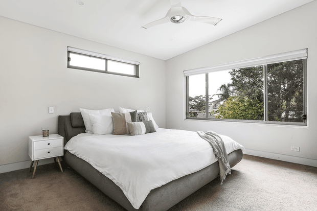 Bright, modern bedroom with a large upholstered bed, white bedding, neutral cushions, a small white bedside table, broad windows overlooking trees, and soft grey carpet. The room has a clean, uncluttered appearance and a contemporary ceiling fan.