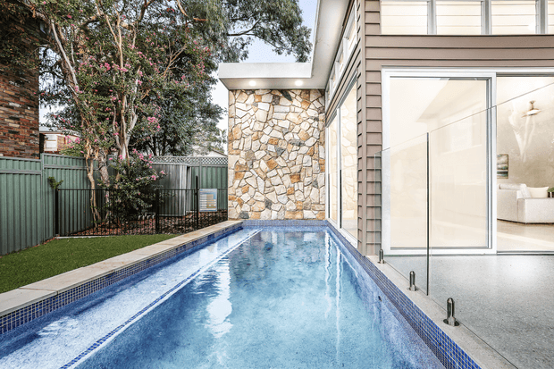 A narrow rectangular swimming pool runs alongside a contemporary home, with blue mosaic pool tiling, pale stone paving, and a glass wall opening into a bright interior. A natural irregular-stone feature wall forms the far end of the pool, while a planted garden and corrugated metal boundary fence occupy the side.