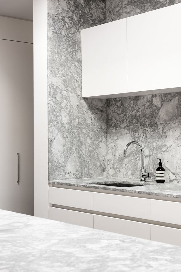 A refined, monochromatic kitchen detail featuring dramatic grey-and-white veined stone wrapping the splashback and benchtop. Flat-panel white overhead and base cabinetry, an undermount sink, and a polished chrome gooseneck tap create a clean, minimalist composition. The foreground appears to be a matching stone island or benchtop.