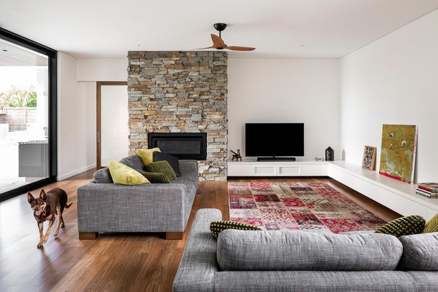 Bright contemporary living room with white walls, warm timber flooring, a prominent stacked-stone fireplace chimney, wall-mounted television, low white built-in media cabinetry, ceiling fan, and two large grey sectional sofas. A colourful patterned rug and yellow-green cushions add contrast, while full-height glazing opens toward an outdoor area.