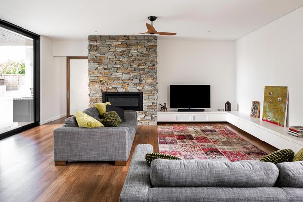 Bright contemporary living room with two large grey sectional sofas arranged around a patterned red rug and a low white media console. A substantial exposed stacked-stone fireplace chimney forms the central feature wall, with a wall-mounted television to the right. Warm timber flooring, white walls and ceiling, large black-framed glazed doors, and a ceiling fan complete the room.