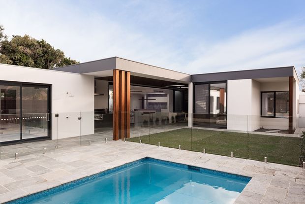 Contemporary single-storey residence viewed from the pool courtyard. The home has a low, flat-roofed composition with crisp white rendered walls, dark-framed floor-to-ceiling glazing, exposed timber-clad structural columns, and an open-plan interior visible through the glass. A paved terrace surrounds the rectangular swimming pool, creating a strong indoor-outdoor connection.