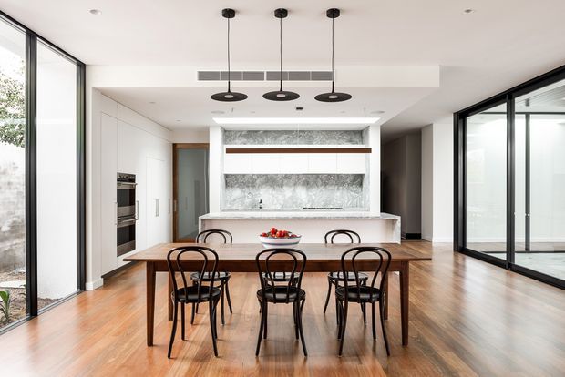 Bright, open-plan kitchen and dining area with white flat-panel cabinetry, a large marble-look splashback, central island, and a timber dining table surrounded by black bentwood-style chairs. Floor-to-ceiling glazing on both sides provides abundant natural light, while three black pendant lights define the dining zone. The space has a clean contemporary appearance with warm timber flooring and strong indoor-outdoor visual connection.
