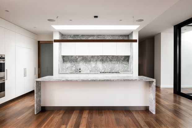 Bright contemporary kitchen centred on a large stone island with waterfall ends and a matching marble-look splashback. Full-height white cabinetry lines the left wall, while a dark timber accent beam and integrated linear ceiling light add definition. Warm timber flooring contrasts with the predominantly white and grey fit-out, and black-framed glazing is visible at right.