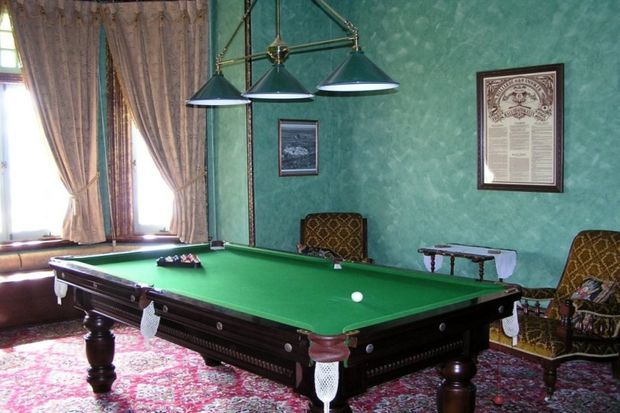 A richly decorated billiards room with a full-size green-felt pool table, dark carved timber detailing, patterned upholstered armchairs, ornate wall artwork, a large Persian-style rug, tall curtained windows, and a three-light suspended fixture. The room has a traditional, historic character with teal textured walls and warm decorative furnishings.