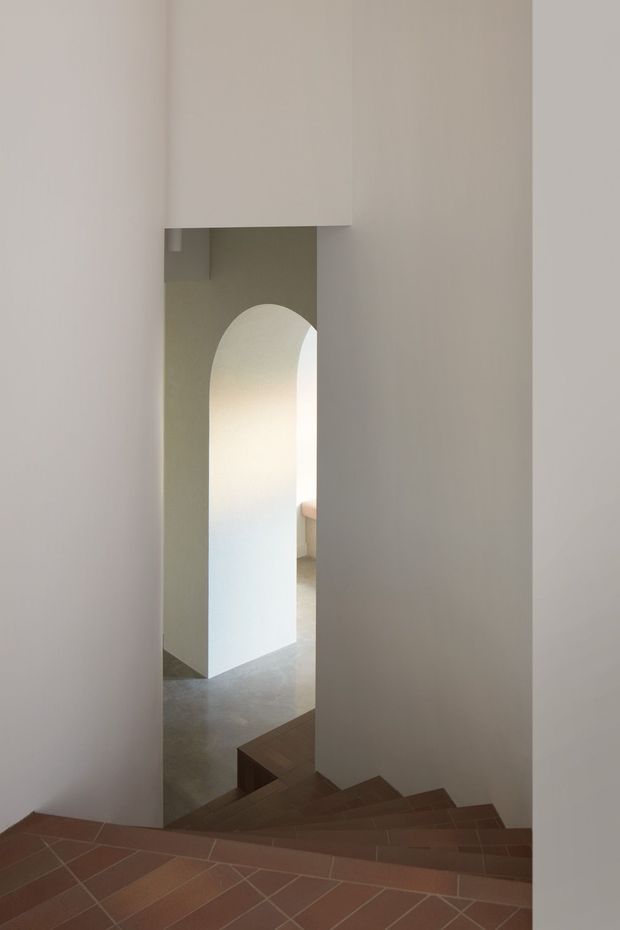 A minimalist interior stairway viewed from an upper landing, with warm terracotta tile flooring, dark timber stair treads, and pale plaster walls. A rounded arch opening leads to a lower-level space, creating a sculptural, softly lit transition between levels.