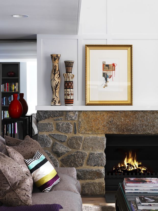 A close-up of a sophisticated living room centred on a rustic irregular-stone fireplace with a black firebox. White decorative wall panelling and a white mantel display framed artwork and tall patterned vessels. A textured upholstered sofa with patterned and colourful cushions sits beside a dark open bookshelf and glass-topped coffee table, creating a refined yet eclectic interior.