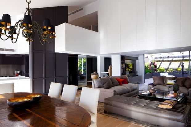Open-plan contemporary interior combining a dining area and living room. A large dark timber dining table with upholstered white chairs sits in the foreground, while grey and brown sofas surround a low coffee table and broad leather ottoman. The space features tall white paneled cabinetry or wall cladding, dark timber built-in elements, large openings to an outdoor area, and an ornate black-and-gold chandelier.
