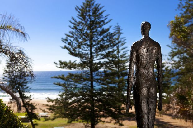 Outdoor coastal scene featuring a tall dark metal human-form sculpture in the foreground, with mature trees, sandy beach, blue ocean, and clear sky visible beyond.