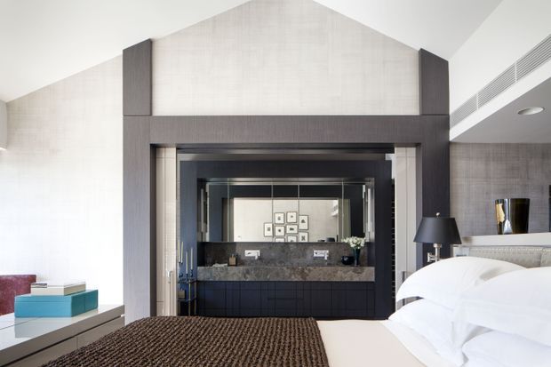 A refined contemporary bedroom viewed toward a dramatic dark-framed opening and mirrored vanity area. The room features a large white upholstered bed with layered white bedding, a textured dark brown bed throw, pale patterned wall surfaces, dark timber or stained-wood architectural framing and cabinetry, integrated linear air-conditioning vents, and a small bedside lamp. The mirror reflects a geometric wall artwork and additional decorative elements, emphasizing the room's depth and symmetry.