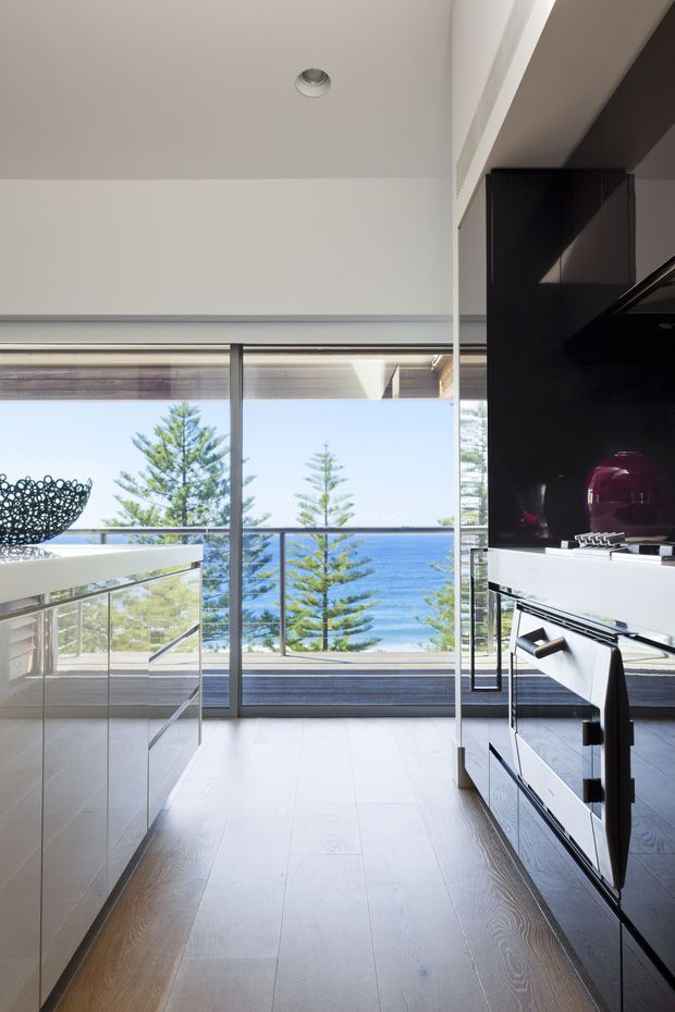 A bright contemporary interior with a large floor-to-ceiling glazed opening overlooking the ocean and coastal trees. The room features pale timber flooring, white walls and ceiling, a sleek dark built-in media or cabinetry wall on the right, and a glass balustrade or partition leading toward an outdoor terrace. Strong natural light and expansive sea views dominate the scene.
