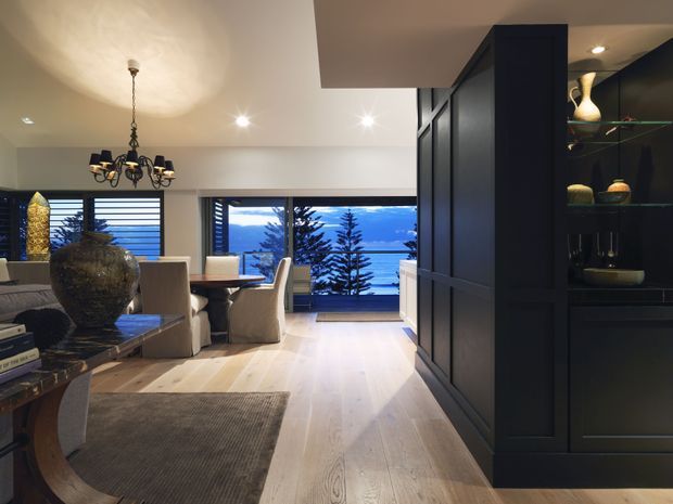 A refined open-plan living and dining area with pale timber flooring, neutral upholstered furniture, a dark built-in display and storage wall, and a traditional chandelier. Large windows and horizontal blinds frame a blue-toned coastal or mountain-like exterior view. The room combines warm wood tones with dramatic dark joinery and soft ambient lighting.