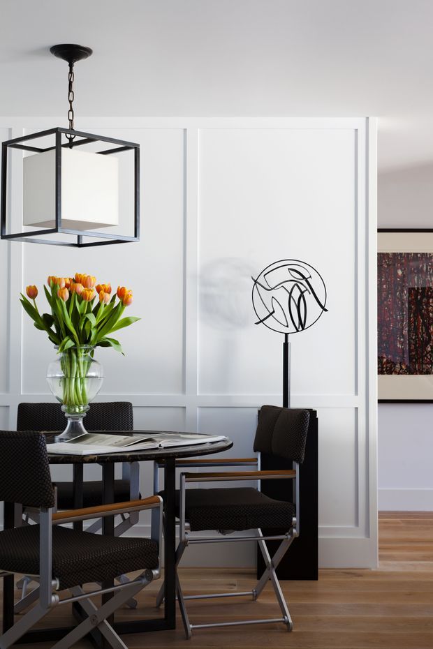 A refined dining area with a round dark dining table, black director-style chairs, a glass vase of orange and yellow tulips, and a geometric black pendant light. White decorative wall panelling provides a bright backdrop, with timber flooring and a framed artwork visible through the adjacent opening. A sculptural black floor lamp adds a contemporary accent.