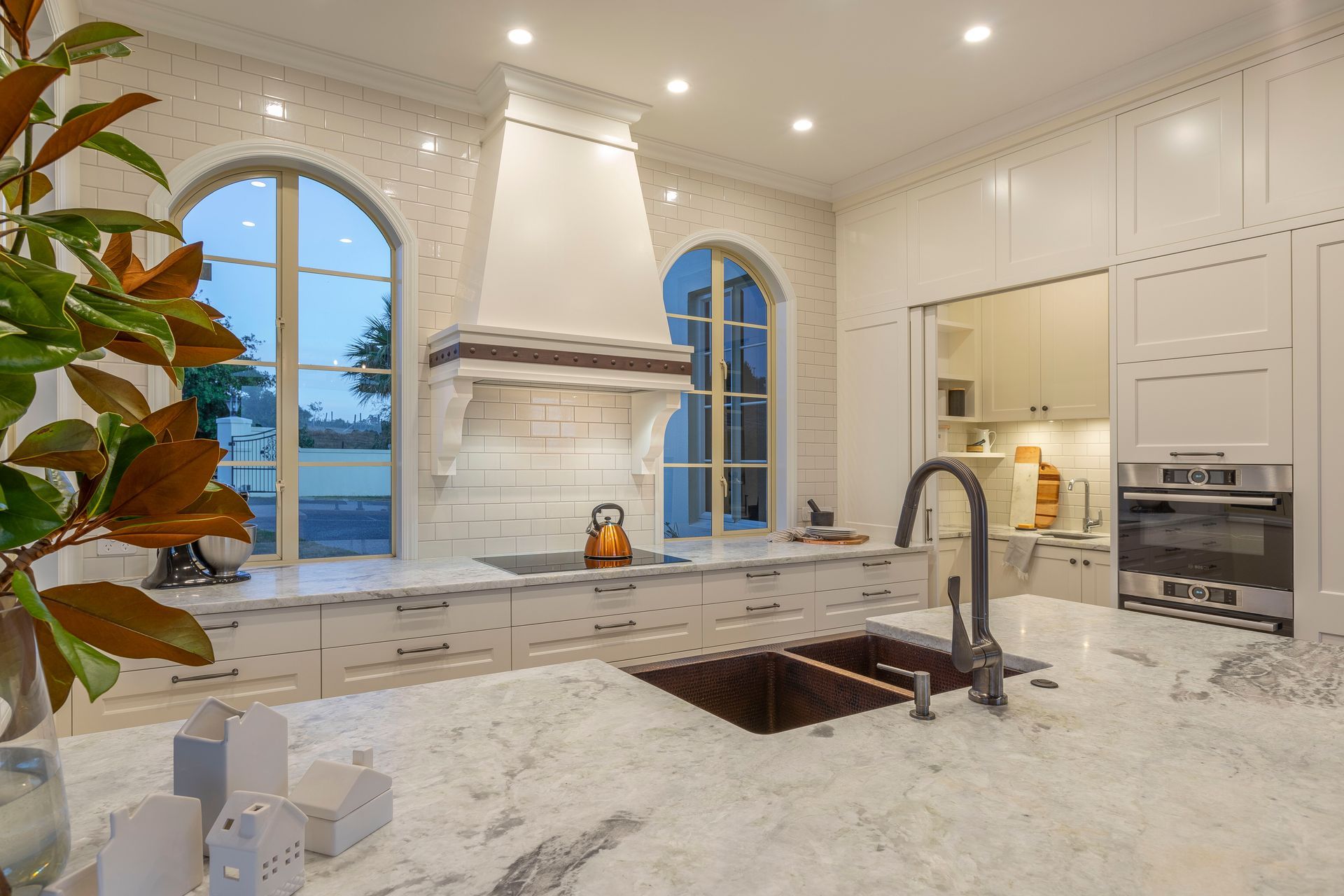 To echo the feature copper sink the clients especially loved, we wrapped a custom-welded then studded copper band to a bespoke rangehood canopy.  It's form is designed to gently rise between two arched windows with a tapered canopy above reminiscent of styles found in French castles. Desiring a scullery, walls were rearranged borrowing from a pool room behind.  Specifying thin sliding-doors optimises seamless flow into the hidden 'room'. Our tiling layout plan ensured no awkwardly small tiles bothered the eye.