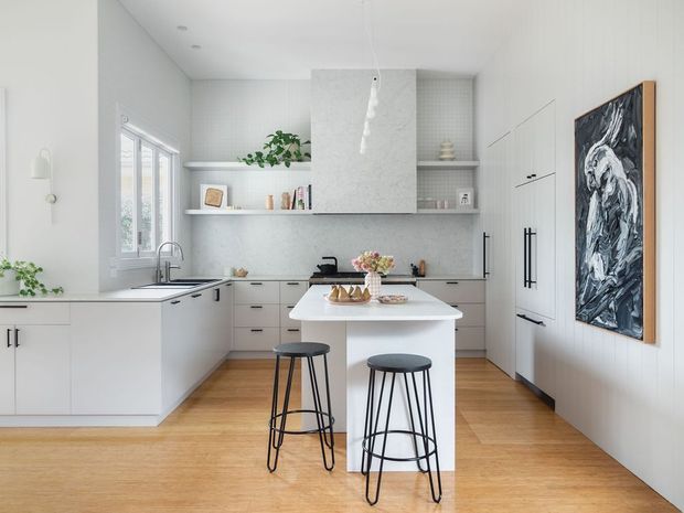 Bright contemporary white kitchen with a central island and two black metal stools. The room features pale timber flooring, white cabinetry, a light stone-look benchtop and splashback, open shelving, integrated appliances, pendant lighting, and a large abstract artwork on the wall.