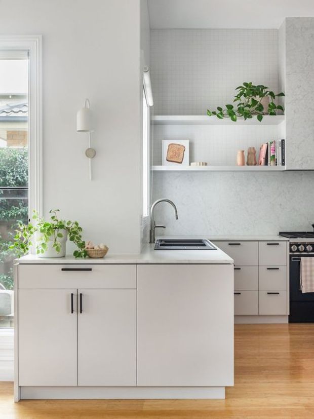 Bright contemporary kitchen with pale cabinetry, a compact island with integrated sink, open shelving, light grey tiled backsplash, pale stone benchtops, timber flooring, and black hardware and appliances. Large window glazing and indoor plants add natural light and greenery.
