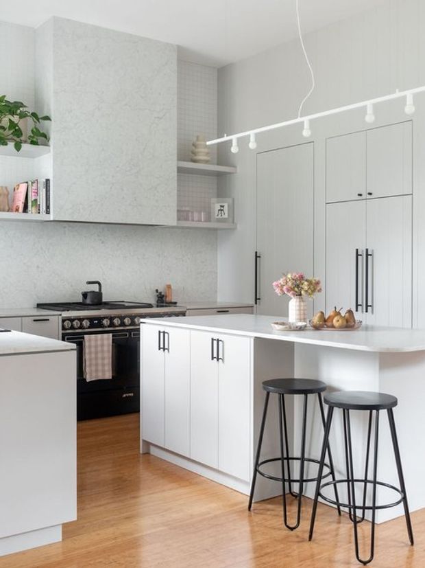 Bright, compact contemporary kitchen with white shaker-style cabinetry, a central island with a raised breakfast bar, black metal bar stools, a freestanding range cooker, pale stone-look surfaces, open shelving, and warm timber flooring. Track lighting and black hardware provide subtle contrast.