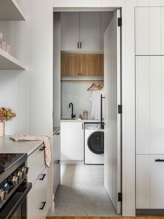 A compact, concealed laundry room viewed through a tall white paneled door within a contemporary kitchen. The laundry includes white cabinetry, warm timber overhead storage, a stone-look benchtop with undermount sink and black tapware, hanging rails, a front-loading washing machine, and pale grey tiled flooring. White kitchen cabinetry and open shelving are visible in the foreground.