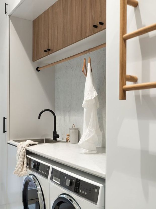 A compact, contemporary laundry room with stacked overhead timber cabinetry, a white benchtop, integrated stainless-steel sink and matte black gooseneck tap. Front-loading washing and drying machines sit beneath the counter. A pale stone-look splashback, timber hanging rail, white garments, and a timber ladder-style wall feature complete the practical, warm-toned fit-out.