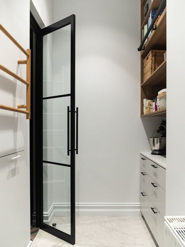 A narrow, bright pantry or utility storage room with white walls, pale tiled flooring, open timber shelving, white drawers and benchtop, integrated appliance storage, and a black-framed glazed door. Timber wall rails provide additional storage on the left.