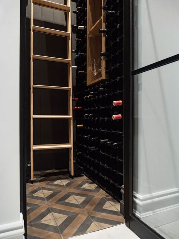A compact wine storage room viewed through an open doorway. The dark walls are fitted with extensive bottle racks holding wine bottles, alongside a tall timber ladder-style shelving unit and a timber display cabinet. The floor features patterned geometric tiles in brown, beige, and dark grey, while the surrounding doorway has dark trim and white decorative wall panelling.