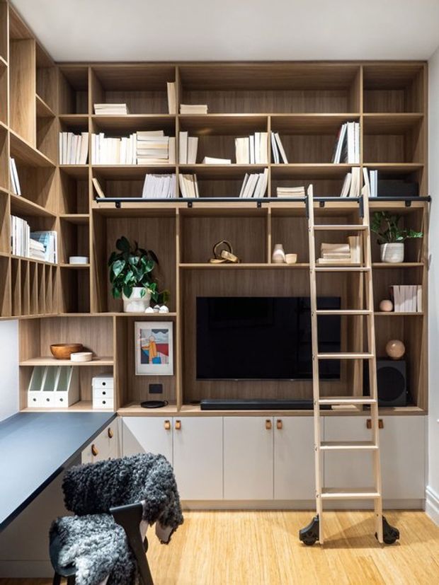 A built-in timber wall unit spans the room, combining extensive open shelving for books and decor with lower white cabinetry, a centrally mounted television, and a rolling library ladder. A dark worktop and desk area occupy the lower left, with a chair in the foreground. The warm wood joinery and light timber floor create a tailored, functional study or home office with a compact media area.