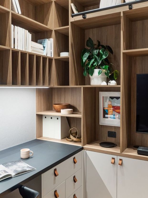 Built-in timber joinery combines open shelving, book storage, a compact desk or work surface, cabinetry, and integrated media storage. The warm wood shelving is styled with books, a houseplant, framed artwork, and decorative objects, while a dark countertop and pale lower cabinets create a refined, contemporary study or living-area fit-out.