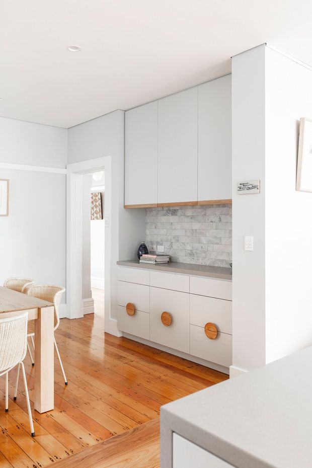 Bright contemporary dining area with a built-in wall of pale grey cabinetry, integrated drawers, warm timber handles, and a grey marble-look tiled splashback. The space has white walls and ceiling, warm polished timber flooring, a light timber dining table with woven white chairs, and a doorway leading to another room. A pale benchtop or island edge is visible in the foreground.