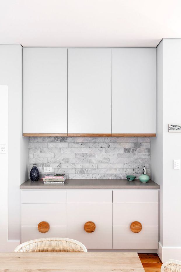 A bright, compact built-in dining or kitchen storage nook with white flat-panel upper and lower cabinetry, warm round timber pulls, a grey stone benchtop, and a horizontal grey-white marble tile splashback. A light timber dining table and woven chairs are partially visible in the foreground, with small decorative objects and books on the counter. The overall design is clean, contemporary, and softly warm.