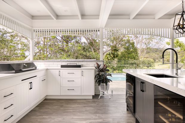 Bright covered outdoor kitchen and entertaining area with white shaker-style cabinetry, a large stone island with integrated sink and black gooseneck tap, built-in barbecue, and a secondary cooking area. The space has a white timber-framed screened enclosure, exposed coffered ceiling beams, grey-brown wood-look flooring, and views toward a swimming pool and leafy garden. A black-framed pendant light and glass-front beverage storage add contrast.