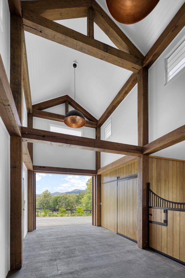 A tall, barn-like interior with exposed dark timber post-and-beam framing, a white vaulted and partially double-height ceiling, concrete flooring, and timber-lined garage or utility doors. Copper pendant lights hang from the structure, while an open glazed doorway frames a landscaped rural mountain view. The space is bright and rustic with strong structural detailing.