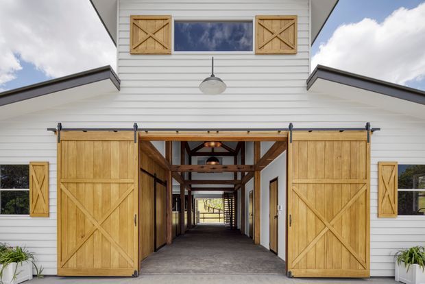 Front-facing view of a distinctive barn-inspired building entrance. White horizontal weatherboard cladding surrounds a large open passage with sliding timber barn doors, matching timber window shutters, exposed dark timber framing, and a concrete floor. The covered corridor extends through the building toward a bright rear opening, with simple industrial-style pendant lights and minimal planting at the edges.