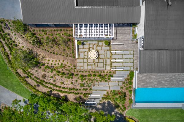 Top-down aerial view of a contemporary property showing dark metal-roofed building wings, a central landscaped courtyard with geometric paving and planted garden beds, a circular feature, timber decking, and a bright blue rectangular swimming pool. The image emphasizes the outdoor site layout and structured landscape design.