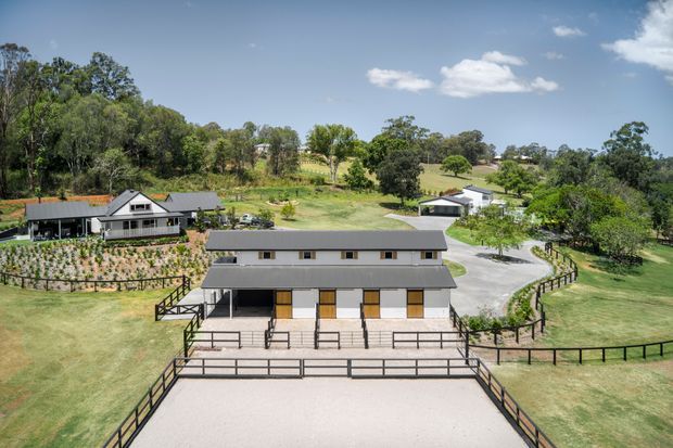 Aerial view of a rural equestrian property featuring a long, modern stable building in the foreground, enclosed paddocks and fencing, landscaped grounds, a secondary house, and a tree-covered gently rolling landscape under a clear sky.