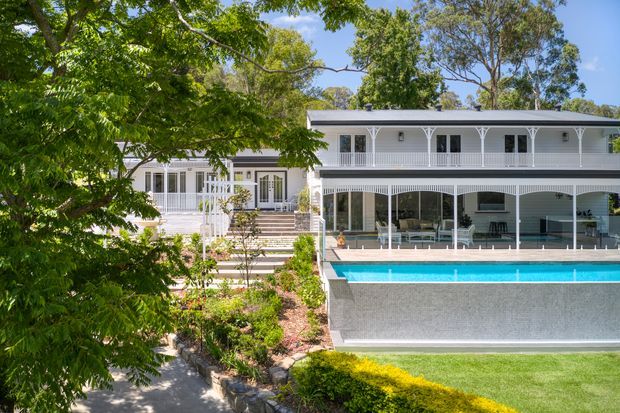 Exterior view of a substantial white two-storey residence set among mature trees and landscaped gardens. The home features a long covered verandah or balcony with slender decorative posts, extensive glazed openings, a low-pitched roof, and a large rectangular swimming pool in the foreground. Steps and pathways connect the upper house level to the landscaped lawn and pool area.