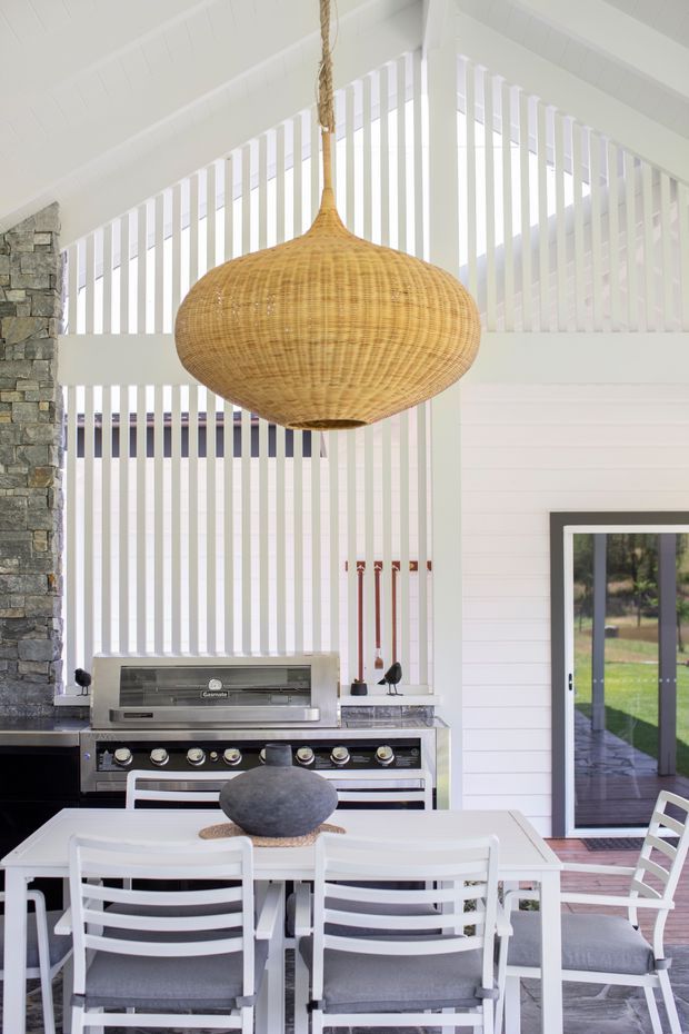 Bright, airy outdoor entertaining area with a built-in barbecue kitchen, white dining table and chairs, a large woven pendant light, and a stone-clad wall. White vertical screening and pitched ceiling elements create a light coastal feel, while sliding doors connect the space to the garden.
