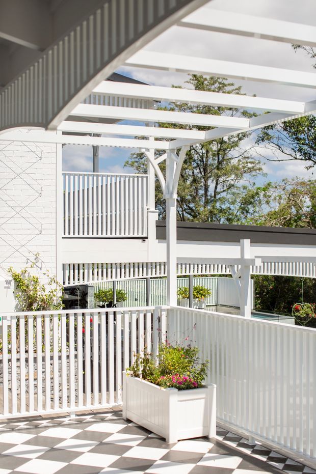 Bright outdoor terrace or courtyard with a white-painted pergola, geometric support posts, extensive vertical balustrading, and a large black-and-white checkerboard tiled floor. White rendered brickwork and planter boxes with flowers frame the space, with trees visible beyond the railing. The overall composition is crisp, airy, and strongly patterned.