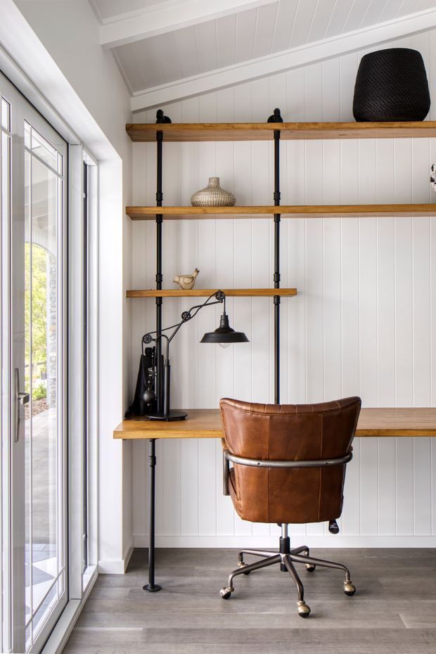 A compact home office nook with a wall-length built-in shelving and desk system. Warm timber shelves are supported by black metal pipe uprights against white vertical wall panelling, with a leather swivel office chair, black articulated desk lamp, decorative objects, and dark grey timber-look flooring. Full-height glazed doors or windows line the left side, bringing in abundant natural light. The ceiling has white painted exposed-style boards and beams.