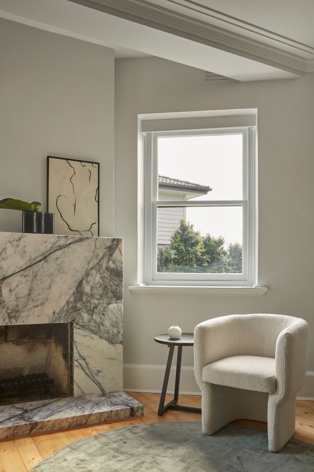 A refined sitting area with a veined marble fireplace, pale neutral walls, detailed decorative ceiling moulding, and a tall white-trimmed window. A sculptural boucle armchair and slim dark side table sit on a muted rug over warm timber flooring, creating a calm, understated interior.