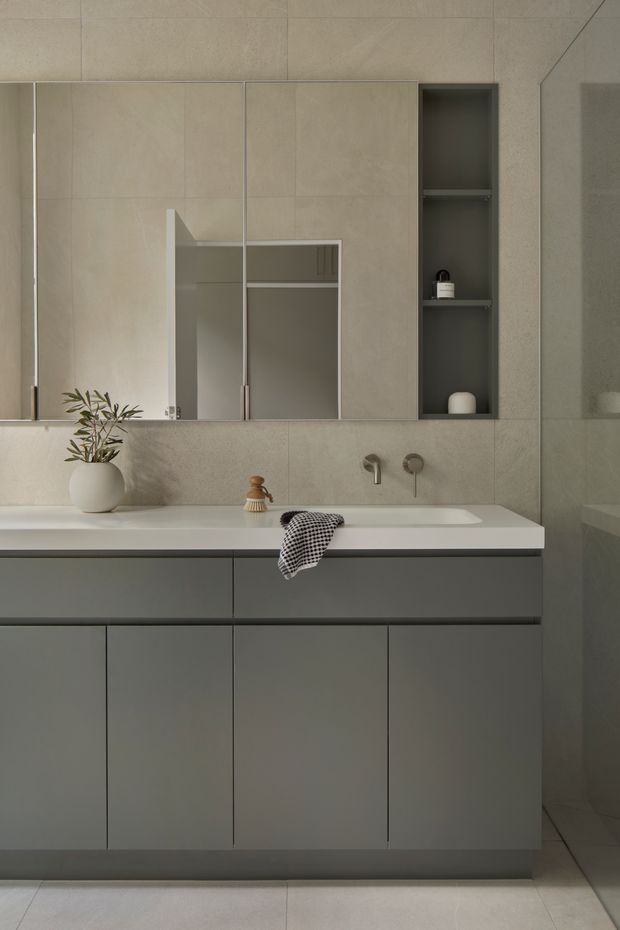 A contemporary minimalist bathroom vanity with a wide pale stone or engineered-stone benchtop, integrated basin, wall-mounted brushed metal tapware, and flat-panel dark grey cabinetry. Large mirrored cabinet doors and an open recessed shelf provide storage above the vanity. The walls and floor are clad in large-format warm grey-beige tiles, creating a restrained, cohesive finish. Small decorative objects and a towel add subtle domestic detail.