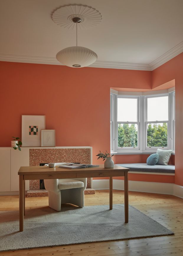 Warm, thoughtfully styled interior featuring a coral-red room with a timber writing desk, upholstered chair, built-in window seat, and a terrazzo-clad fireplace surround. A projecting bay window brings in natural light, while ornate ceiling cornices and decorative ceiling roses support the period character. Pale timber floorboards, a neutral rug, framed artwork, and sculptural pendant lighting complete the inviting workspace.