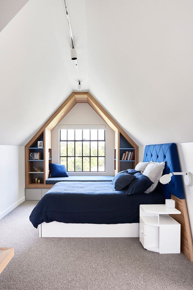 A bright attic bedroom with a steeply pitched, gabled ceiling and white walls. A large bed is dressed in deep navy bedding and has a striking blue upholstered headboard. Built-in timber shelving and cabinetry frame the gable-end window, creating compact storage niches. The room features a white bedside unit, contemporary wall-mounted reading lights, and a neutral grey carpet.