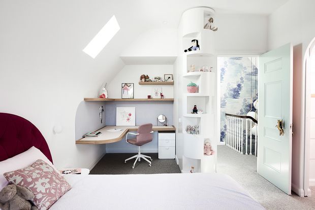 Bright, compact bedroom with a built-in study nook beneath a sloped ceiling. The room features a curved burgundy upholstered bedhead, pale bedding, a timber desk with white cabinetry, a mauve desk chair, open shelving, and a tall curved display unit. A pastel blue door opens to a hallway or landing with a visible stair balustrade and patterned wallpaper. The overall look is playful, clean, and softly coloured.