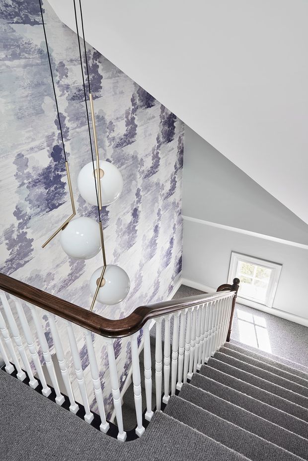 A bright residential stairway viewed from an upper landing, featuring grey carpeted stairs, white turned balusters, and a dark polished timber handrail. A tall stairwell wall is covered in a pale lavender and grey scenic patterned wallpaper, complemented by a sculptural multi-globe pendant light with slim dark and brass-toned arms. White trim, grey walls, and a small window complete the refined traditional interior.