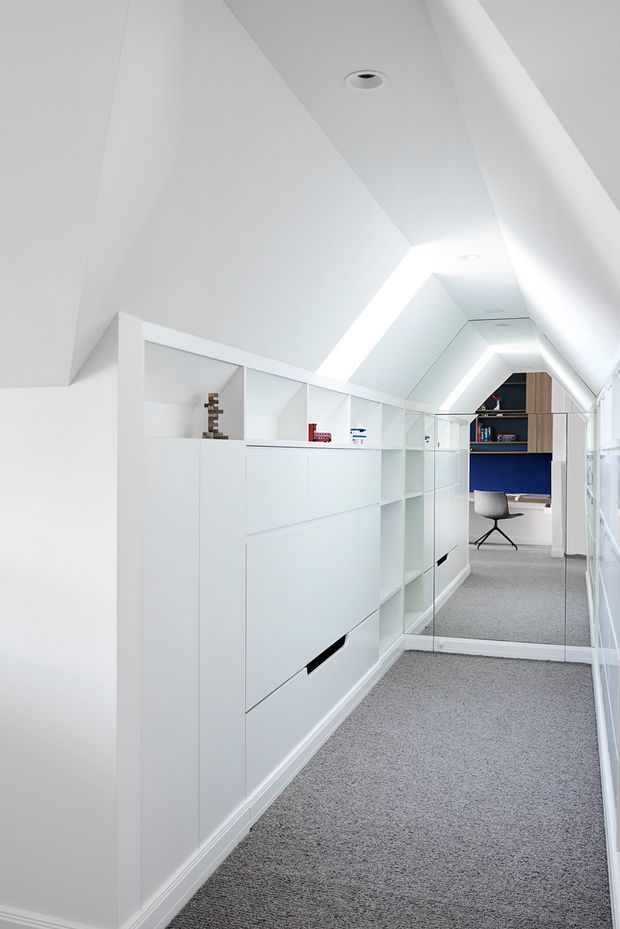 A bright, narrow attic workspace or study with a sharply sloped white ceiling, recessed downlights, integrated white storage cabinetry and open shelving, and grey carpet flooring. A mirrored panel enlarges the corridor-like space and reflects a compact desk area with a blue feature wall, timber shelving, and a modern task chair. The design is clean, highly built-in, and minimalist.