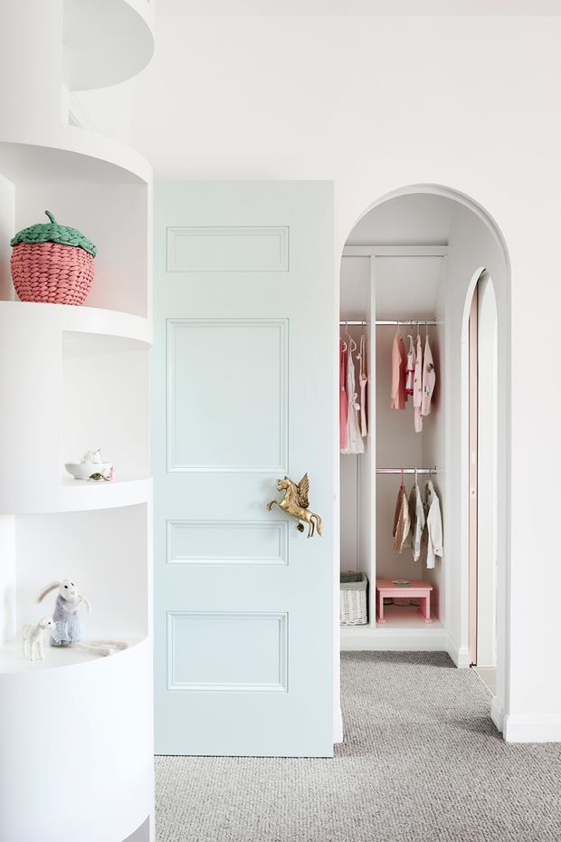 Bright, playful children's dressing area with a pale blue paneled door, ornate gold horse-shaped handle, curved white built-in shelving displaying toys and decor, and an arched opening into a compact wardrobe filled with children's clothing. The space has white walls and joinery, soft pink accents, and grey carpet flooring.