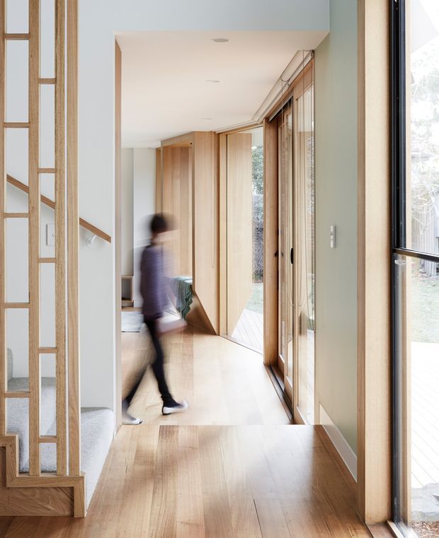 A bright residential hallway with warm timber flooring, extensive light-wood built-in joinery, and full-height timber-framed glazing along the right side. A timber-framed decorative screen and stair opening are visible on the left, while a blurred person walks through the corridor. The space has a clean, contemporary character with a long sightline and abundant natural light.