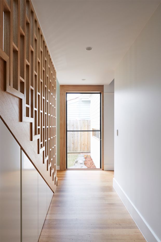 A bright, minimalist residential hallway viewed beside a staircase. The space features pale timber flooring, white walls and ceiling, a sculptural timber stair with slim metal or glass balustrade, and a full-height timber slatted screen forming a striking geometric partition. At the end of the hall, a black-framed glazed door opens toward a small enclosed outdoor area.